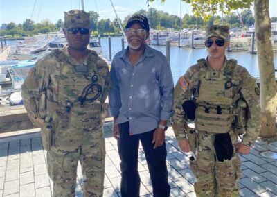 Simeon Fowler standing between two Guardsmen, outside under a shady tree with smaller boats docked in the waterway behind them.