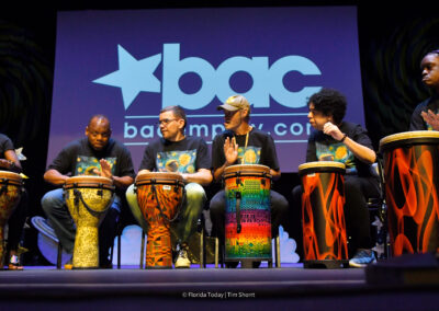 Five performers from the BAC Dynamites sitting beside teaching artist Dallas Clark drumming African beats.