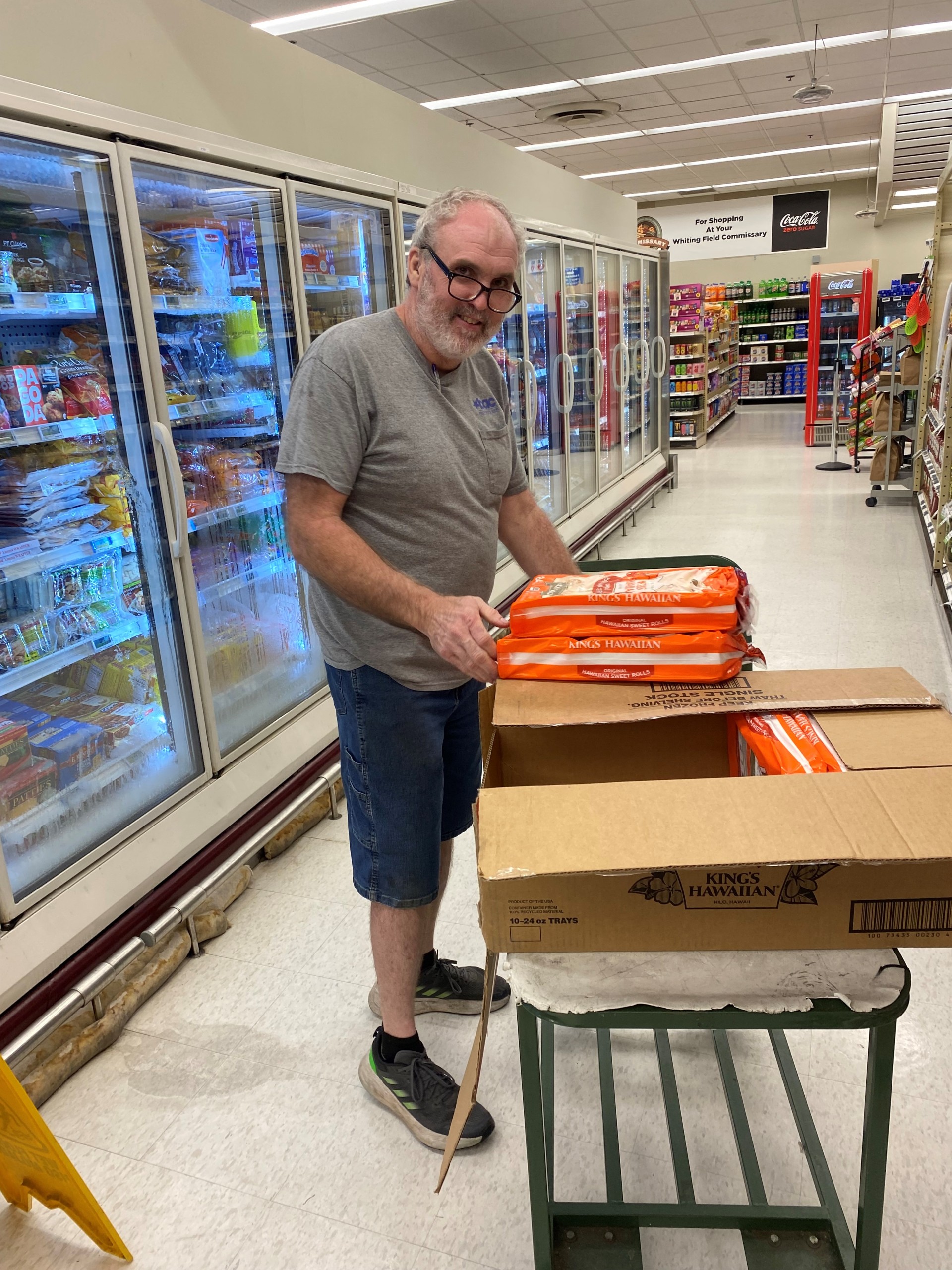 Image: BAC employee Bobby Cleckler wearing a grey BAC work t-shirt and jean shorts. He is smiling and wearing black framed corrective eyeglasses and grey athletic shoes. He stands next to a freezer at a grocery store, commissary, at Whiting field. He is pulling packages of Hawaiian rolls out of a cardboard box which is propped on a wheeled metal stocking cart.