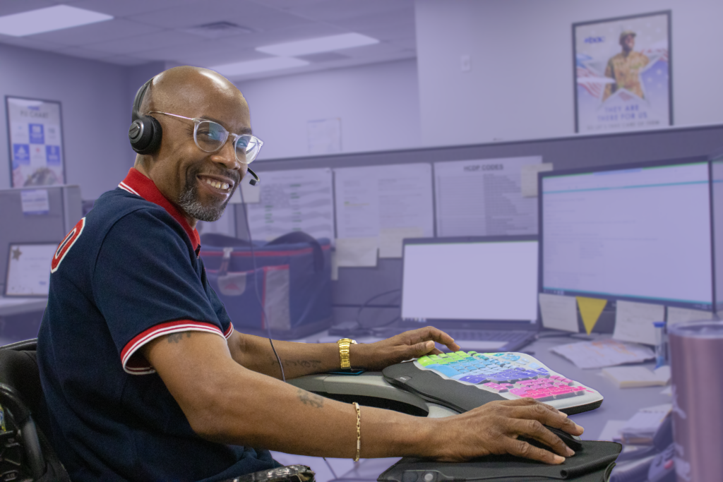 A smiling black man with a headset on sits at a desk with his hand on a computer mouse